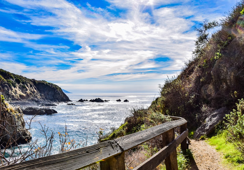 A trail lined with wooden railing leading to ocean cove under bright blue sky streaked with white clouds.