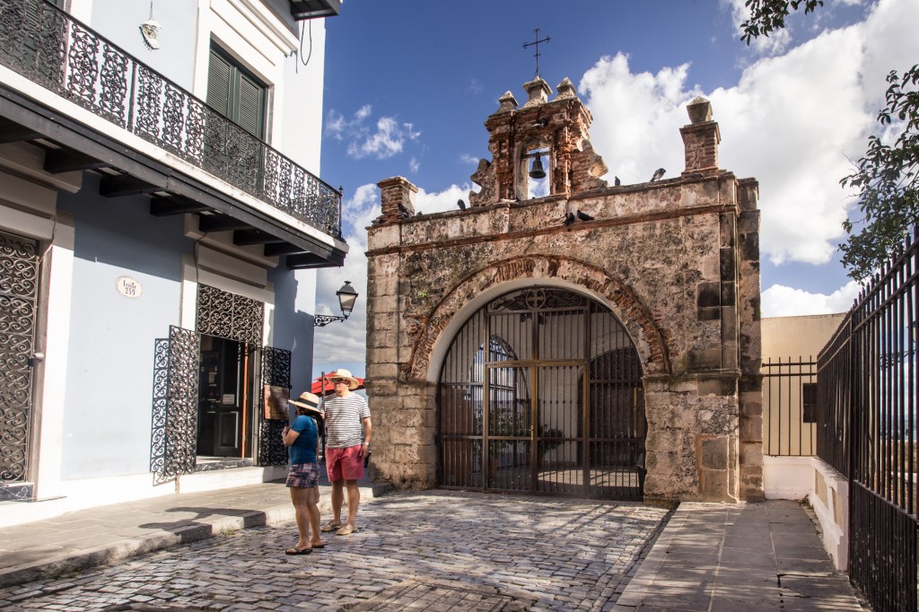 An ornate arched gate made out of stone at the end of a cobblestone street with two people in front of it.